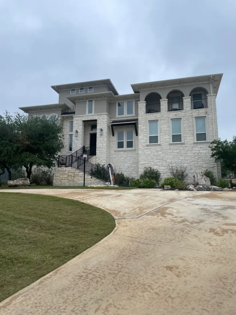 Large modern house with light stone exterior, black trim, and balconies, stained driveway, curved pathway, surrounded by greenery, overcast sky in background