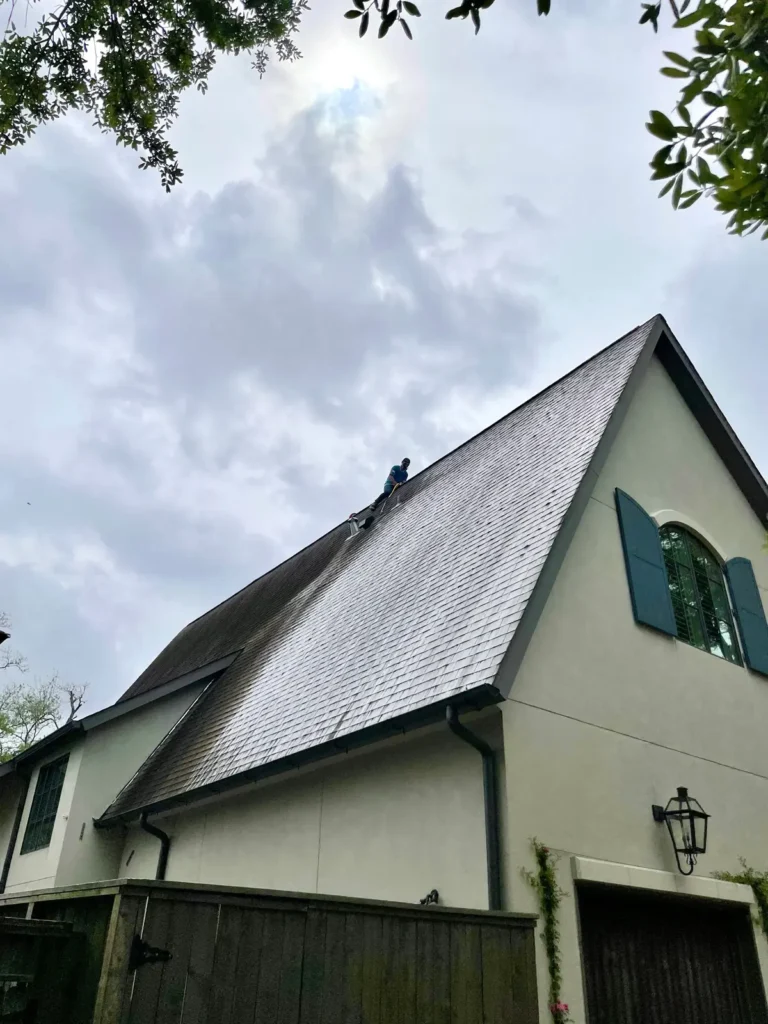 Worker on steep house roof using pressure washer to clean dark stains, partially cleaned section visible, cloudy sky and greenery in background