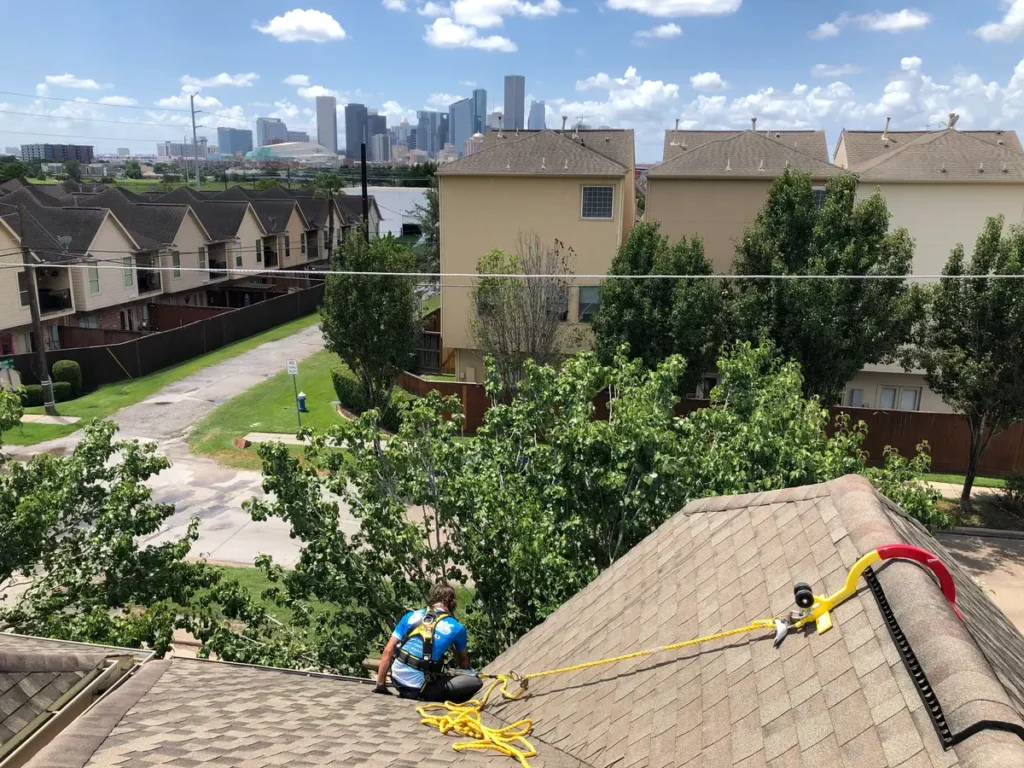 Worker performing roof and gutter maintenance on a shingled rooftop using safety harness and rope anchor with city skyline and residential houses in the background