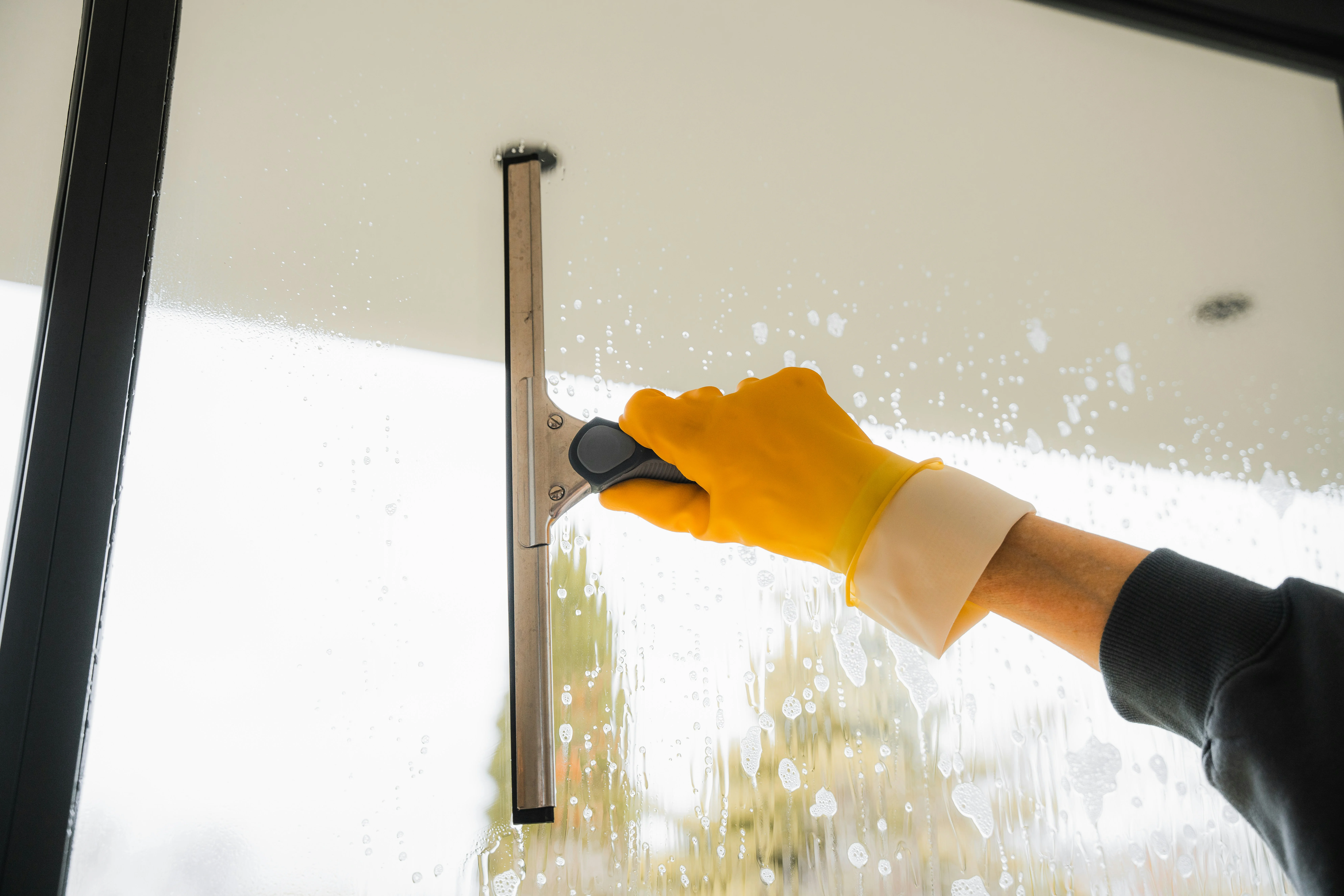A hand in a yellow glove cleaning a hard water stain on window.