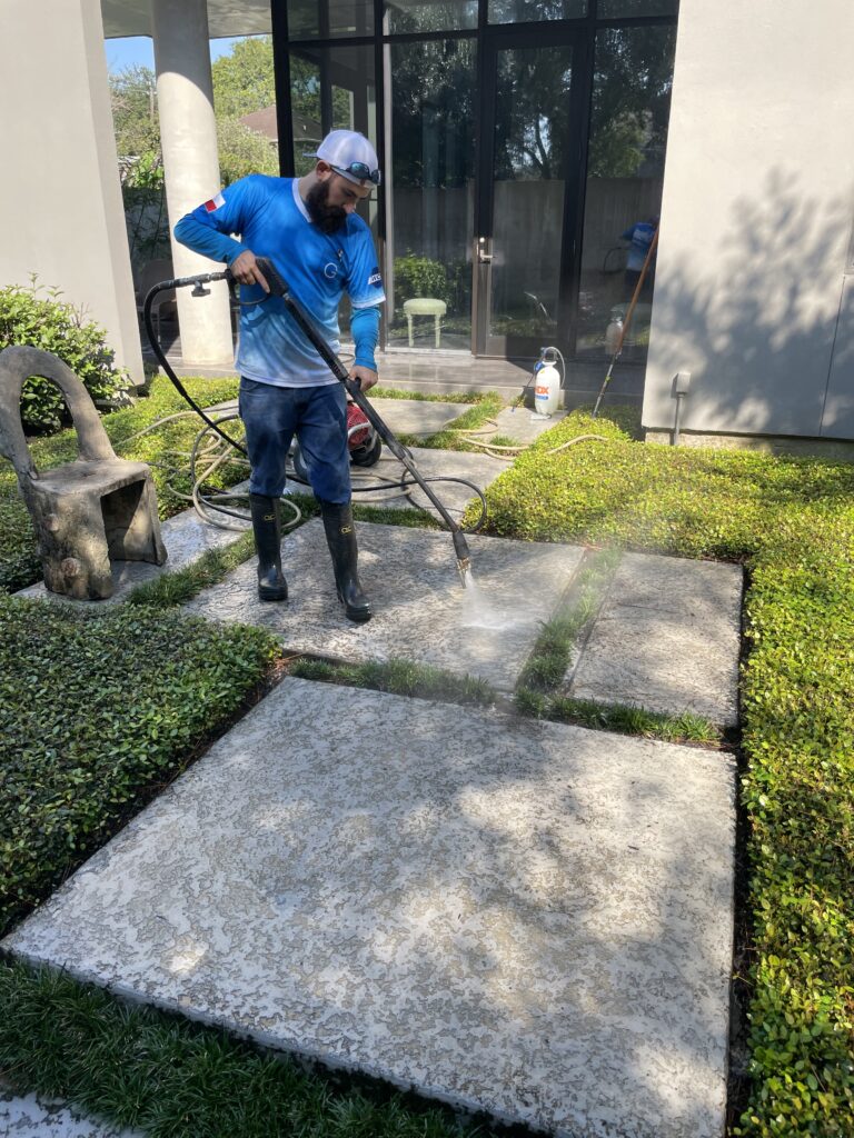 Worker in blue shirt using pressure washer on outdoor concrete pathway surrounded by greenery and modern house with large glass windows in background