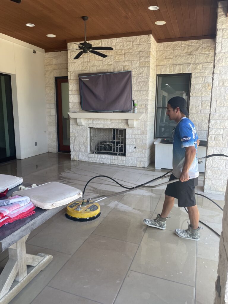 Worker using surface cleaner for pressure washing deck on covered patio with stone fireplace and ceiling fan removing dirt and grime from tiled floor