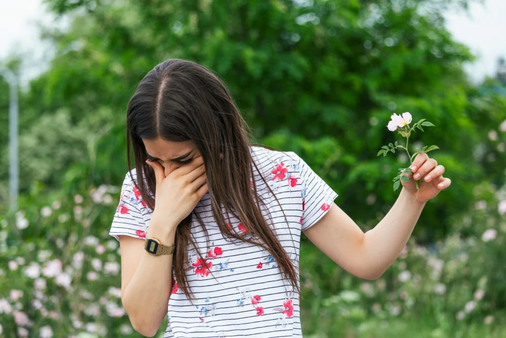 A girl turning her head away while she sneezes near a flower because she doesn't know best time to open windows during allergy season.