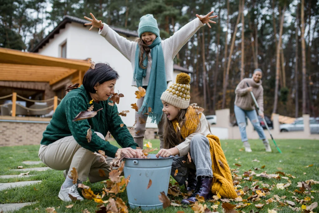 A group of two young girls and an older women playing with fall leaves while doing seasonal home care tasks.