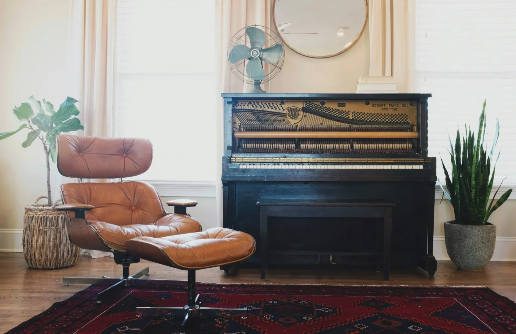 A piano in a room beside a orange chair surrounded by windows on either side.