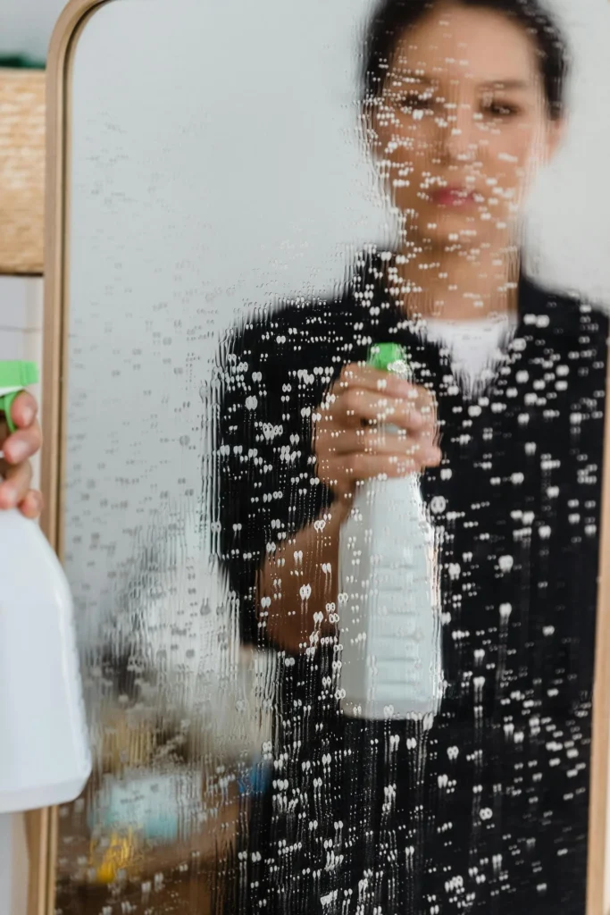 A woman holding up a spray bottle to a glass mirror.