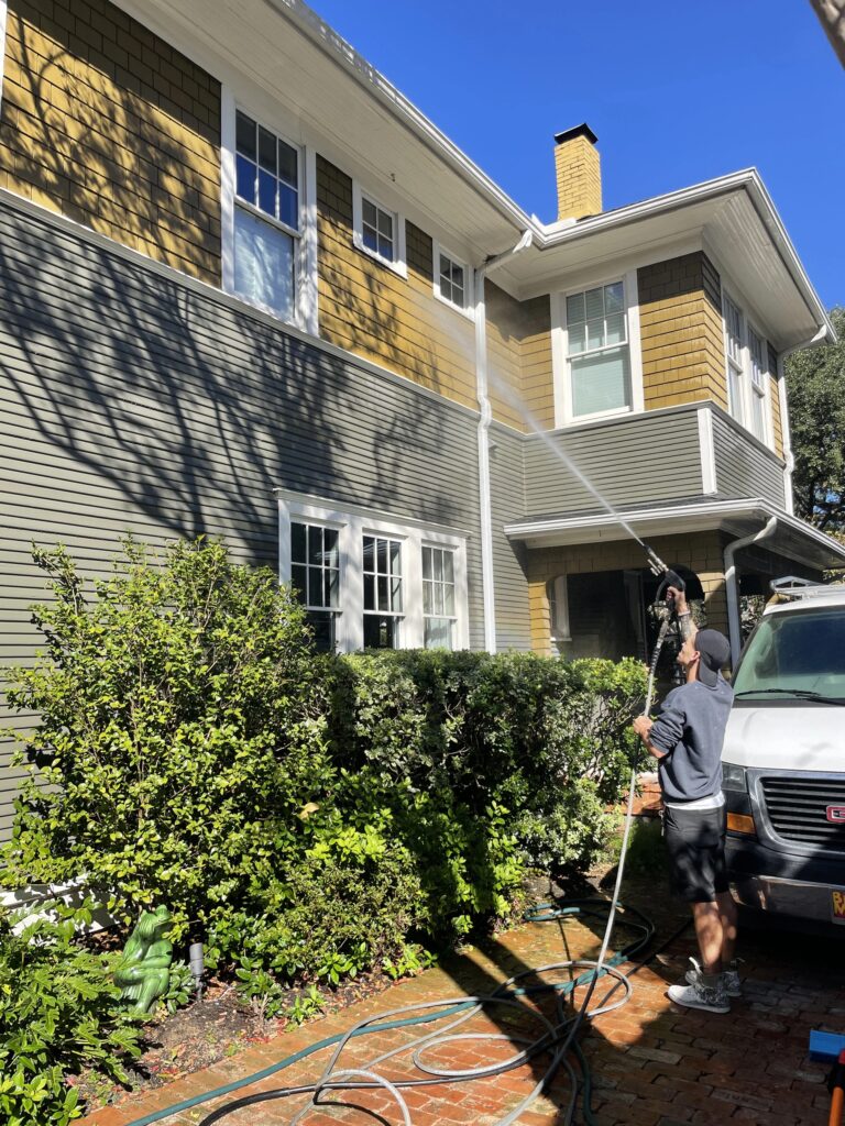 Man aiming pressure washer hose towards second floor of home and cleaning windows