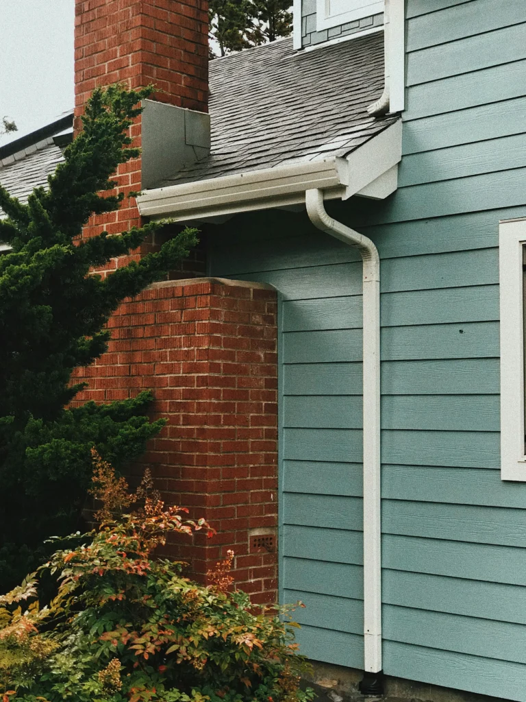 Close up of a house corner with brick chimney blue siding white gutters and greenery scaled