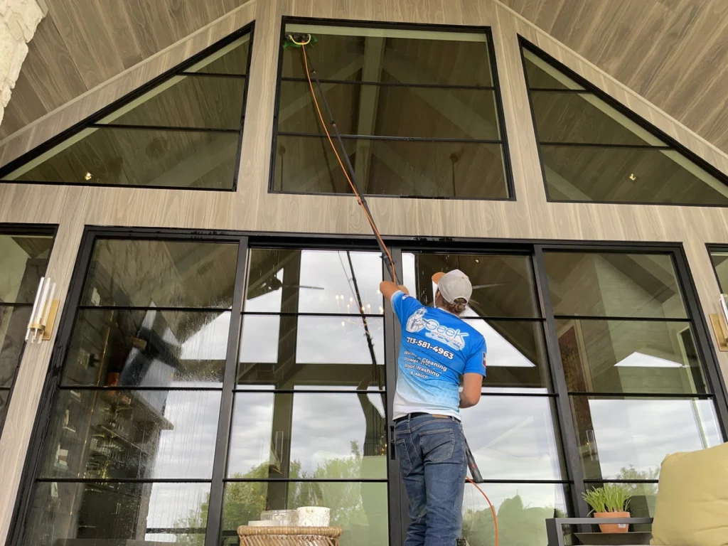 Worker in blue shirt cleaning large glass windows using a water fed pole on modern house exterior